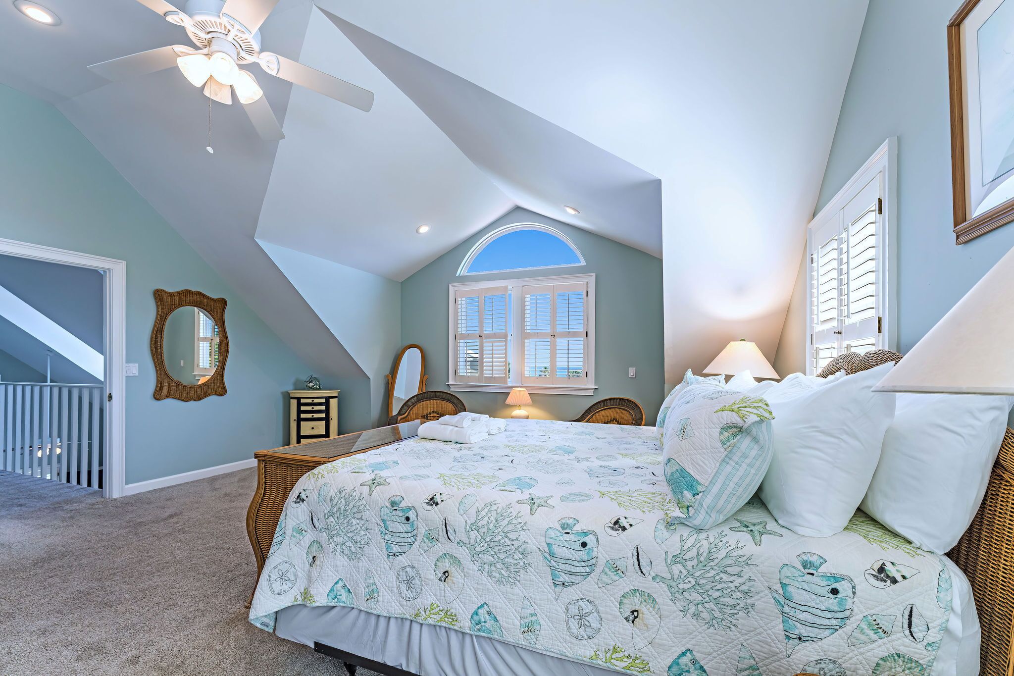 Une chambre avec un plafond en pente, un grand lit recouvert d’une courtepointe matelassée, une fenêtre avec des volets et un ventilateur de plafond.