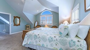 Una camera da letto con soffitto spiovente, un letto grande con copriletto trapuntato, una finestra con persiane e una ventola a soffitto.