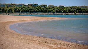Une plage à l’eau claire, avec un rivage sablonneux et des maisons en arrière-plan.