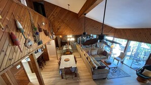 Un vaste espace de vie aménagé dans un style « loft », avec un plafond haut, des poutres en bois, une table à manger, des canapés et une grande fenêtre donnant sur l’extérieur.