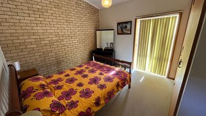 Une chambre avec un mur en briques, un lit recouvert d’une couvre-lit à motifs floraux, une table de chevet, une commode, un miroir et une fenêtre avec des rideaux.