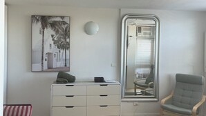 Un espacio habitable pequeño con un aparador blanco, un cuadro enmarco con una escena costera, un espejo grande, una silla verde y una mesa de madera.
