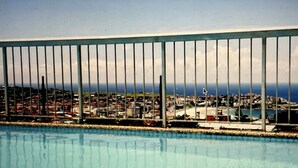 Una piscina con vista a un paisaje urbano costero, una baranda metálica y un cielo despejado.