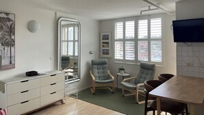 Una habitación moderna con un espejo grande, una mesa de comedor, dos sillones, una pequeña mesita auxiliar y un televisor montado en la pared.