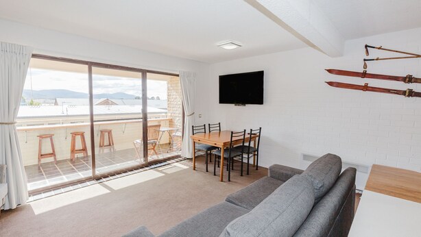 A modern living room with a sofa, a dining table with chairs, a TV mounted on the wall, and a balcony with a view of the cityscape. - Central Park - 10/1 Snowy River Avenue (Jindabyne)