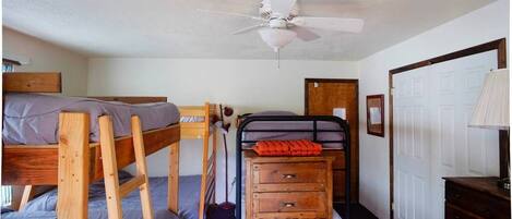 A dormitory room with bunk beds, a ceiling fan, a wooden dresser, and a door.
