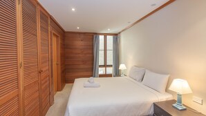 A bedroom with a wooden wardrobe, a bed with white linens, two bedside lamps, and a window with curtains.