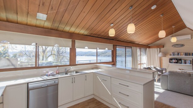 A modern kitchen with a large island, wooden ceiling, and large windows overlooking a lake.