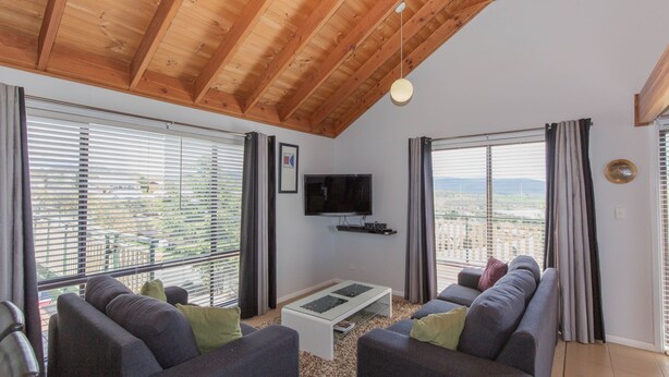 Ein Wohnzimmer mit Holzbalkendecke, großen Fenstern, einem Flachbildfernseher, einem Couchtisch und einer Couch mit Kissen.