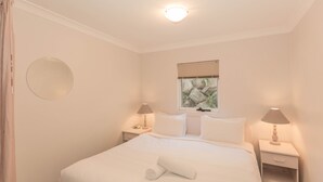 A neatly made bed with white linens and a rolled towel, two bedside lamps, a round mirror on the wall, and a window with a view of rocks. - Horizons - 505/10 Kosciuszko (Jindabyne)