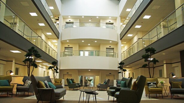 Eine geräumige Lobby mit mehreren Sitzbereichen, einer zentralen Treppe und einer Glasbrüstung im oberen Bereich.