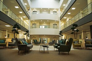 Een ruime lobby met meerdere zithoeken, een centrale trap en een glazen balustrade op de bovenverdieping.