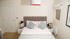 Ein ordentlich eingerichtetes Schlafzimmer mit einem großen Bett, zwei Nachttischen, einem Beistelltisch, einer Lampe und einem Fenster mit Jalousie.