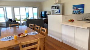 A dining area with a table set for breakfast, a kitchen with a fruit bowl, and a view of the sea through large windows.