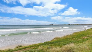 A beach with waves, grassy areas, and a distant shoreline with trees and a lighthouse.