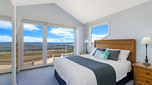 A bedroom with a large bed, a nightstand, a lamp, and a view of the beach through a sliding glass door. - A Top Deck - vast views of the Southern Ocean (Port Fairy)