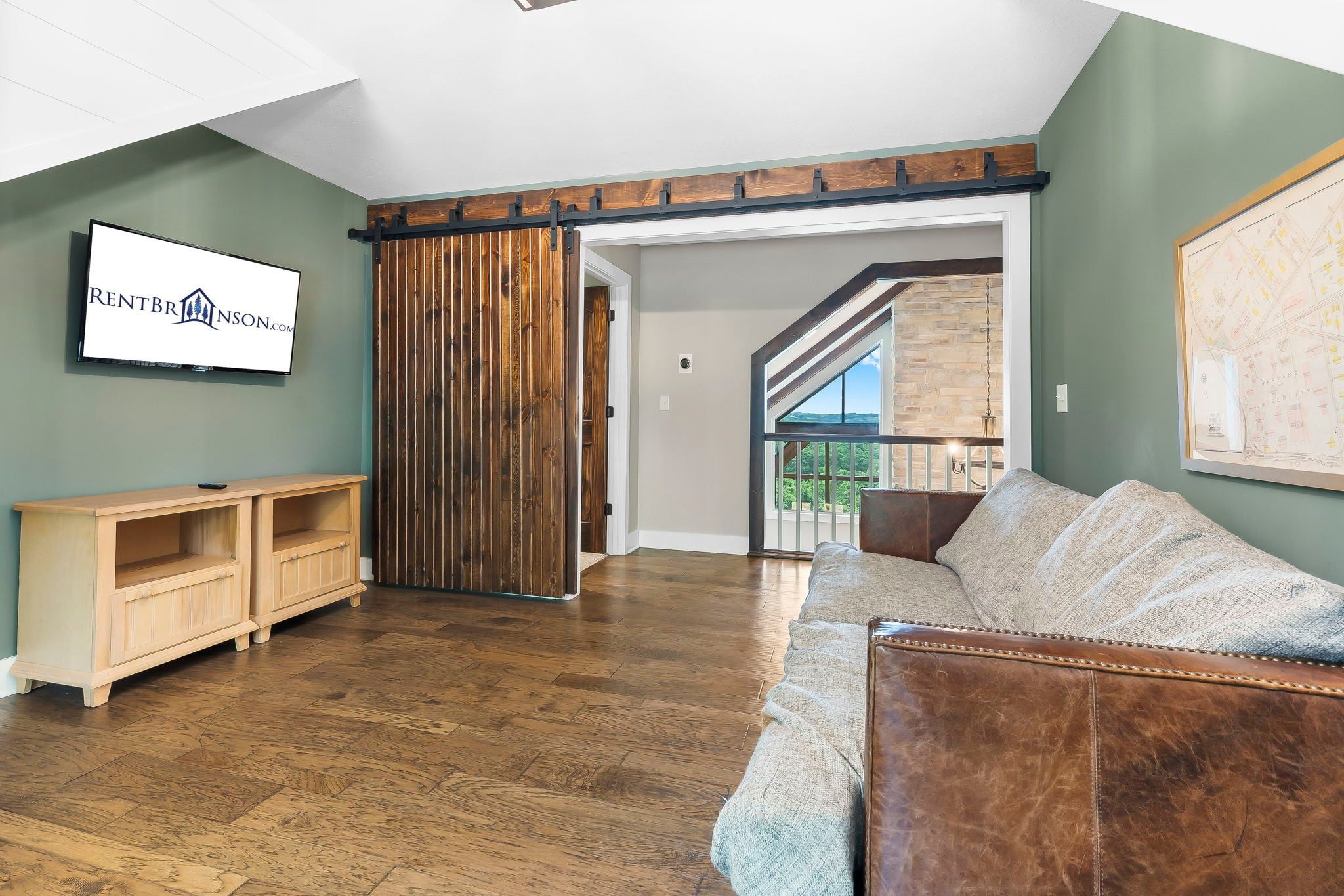 A spacious living room with a wooden floor, a sliding barn door, a mounted television, and a framed map on the wall.