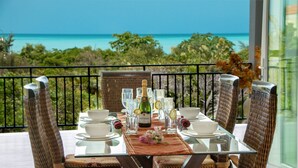 Une table à manger dressée avec de la verrerie, une bouteille et un arrangement floral au centre, sur un balcon donnant sur une mer turquoise.