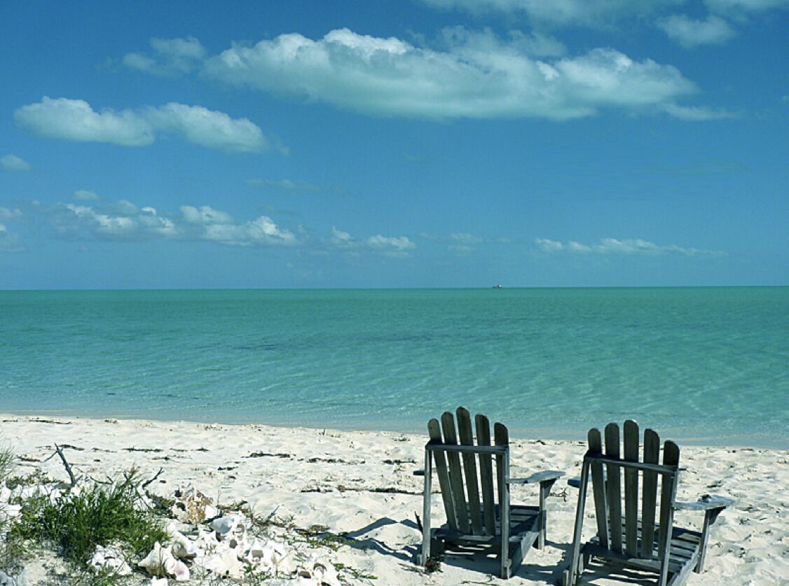 Two Adirondack chairs on a sandy beach with clear turquoise water and a blue sky.