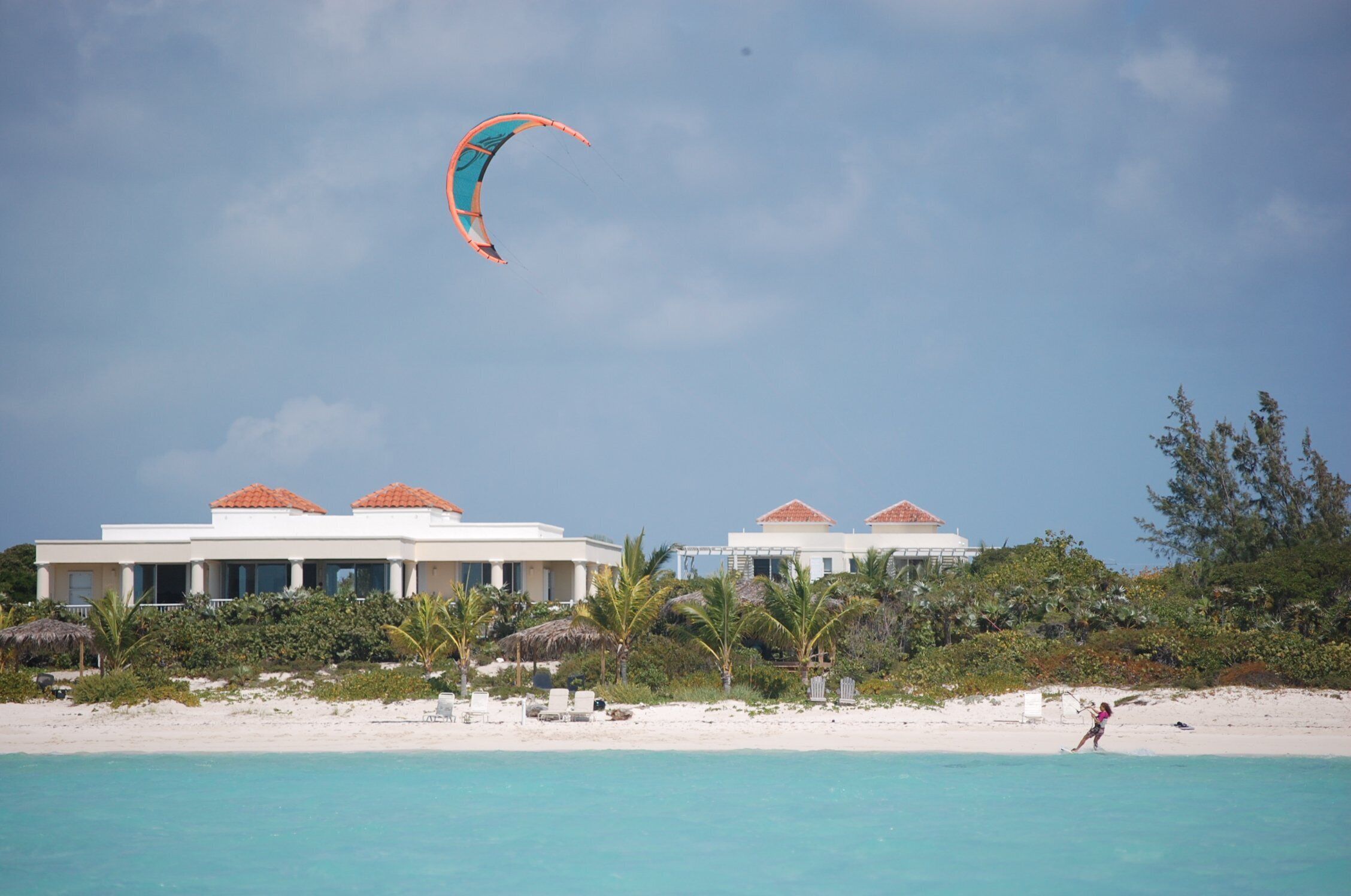 Un complexe hôtelier situé en bord de mer, composé de plusieurs bâtiments, avec un ciel dégagé et une personne pratiquant le kitesurf.