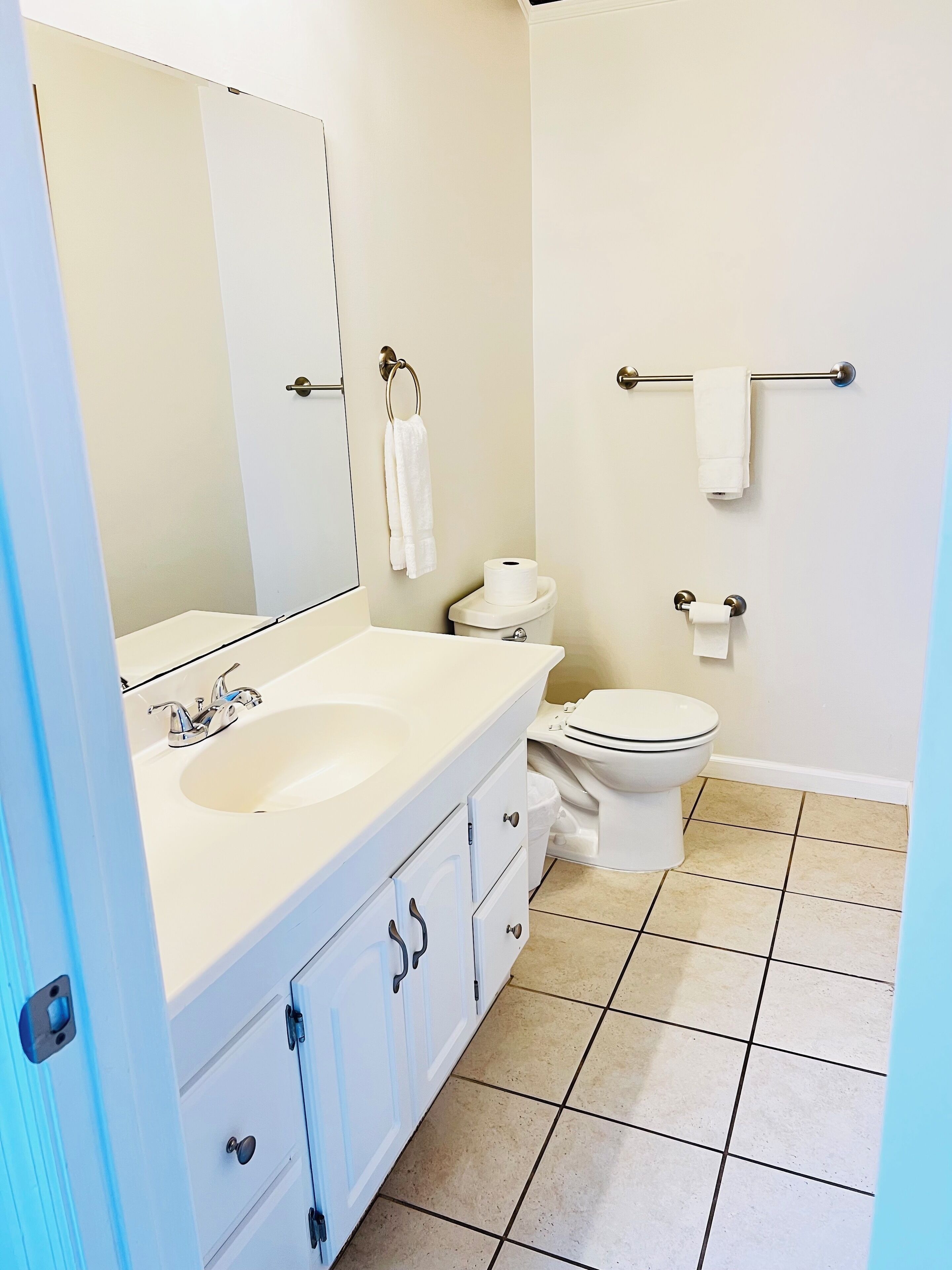 A bathroom with a white sink, mirror, and toilet. There are towels hanging on a rack and a roll of toilet paper on the wall.