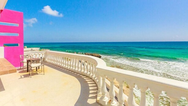 A balcony with a view of the ocean, a white railing, and a pink wall.