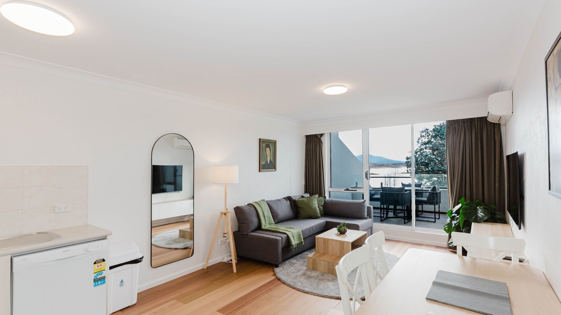 A modern, well-lit living room with a sofa, a coffee table, a TV, and a large window offering a view of the outdoors.