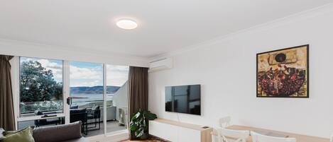 A modern living room with a large TV, a dining area, and a view of a lake through large sliding doors.