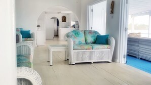 A spacious living area with a white sofa, a white armchair, and a coffee table. There is a kitchen visible in the background, and a large window provides natural light. - A Surfer's Home Away From Home! Soft round the edges, cozy and beachy (Oistins)