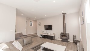 A modern living room with a sofa, TV, entertainment unit, and a wood-burning stove. - 43A Twynam Street - Spacious four bedroom home in central Jindabyne location (Jindabyne)