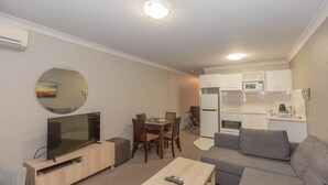 A modern living room with a flat-screen TV, a dining area with a table and chairs, a gray sofa, and a kitchenette with white cabinets and appliances.