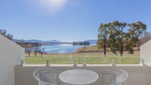 A balcony with two metal chairs and a table overlooking a lake and distant mountains.
