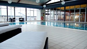 An indoor swimming pool with tiled flooring, lounge chairs, and a view of the outside through large windows.