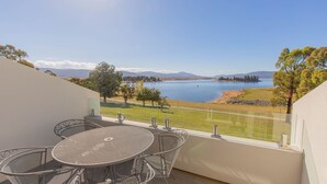 A balcony with a round metal table and chairs, overlooking a lake with mountains in the distance.