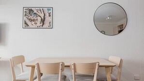 A dining area with a light wooden table and four chairs, a framed picture on the wall, and a round mirror.