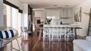 A modern kitchen with white cabinets, a dining area with white chairs, and a wicker chair with a striped cushion. - Spinnakers Waterfront (Port Fairy)