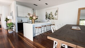 A modern kitchen with white cabinetry, a central island with stools, and a dining area with a dark wooden table. - Spinnakers Waterfront (Port Fairy)