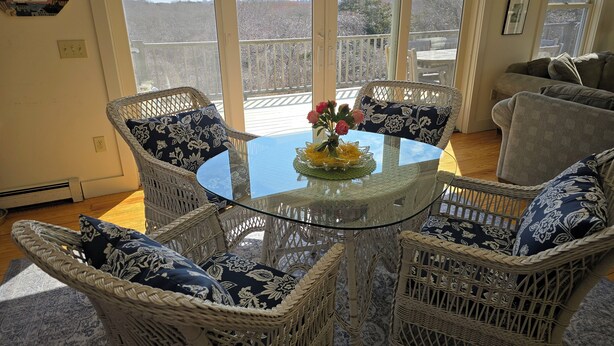 A room with wicker chairs and a glass table, a view through large windows, and a wooden deck outside.