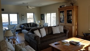 A living room with a sofa, a coffee table, and a bookshelf. There are windows and a ceiling fan.
