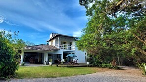 A two-story house with a covered patio, surrounded by lush greenery and trees. - Casa Quinta da Margem (Florianópolis)
