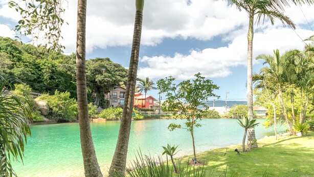 A tropical landscape with a clear lake, palm trees, and a house in the background. - Casa Quinta da Margem (Florianópolis)