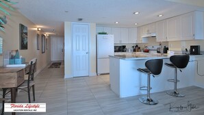 A modern kitchen with white cabinets, a central island with bar stools, and a dining area with a wooden table and chairs. - Chateaux Condominium 404-Breathtaking gulf views! (Indian Shores)