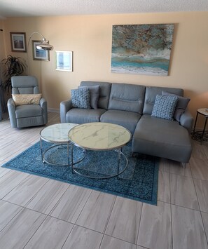A living room with a grey sofa, two armchairs, a glass coffee table, and a blue patterned rug. - Chateaux Condominium 404-Breathtaking gulf views! (Indian Shores)