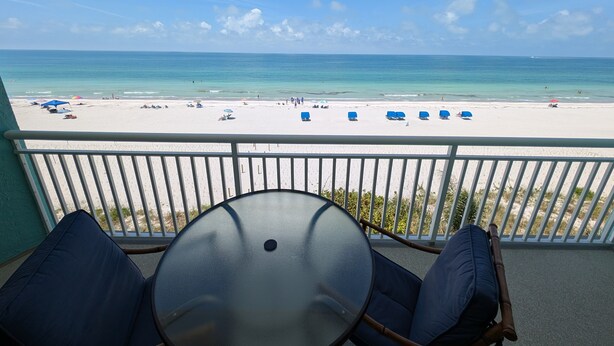 A balcony with a glass table and chairs, overlooking a beach with several sun loungers and people. - Chateaux Condominium 404-Breathtaking gulf views! (Indian Shores)