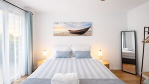 Une chambre d’hôtel avec un lit, un ventilateur de plafond, un tableau représentant un bateau sur la plage, une table de chevet avec une lampe et un miroir au mur.