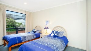 A bedroom with two beds, a nightstand with a lamp, a window with curtains, and a view of greenery outside. - Million Dollar Sea Views (Hayborough)