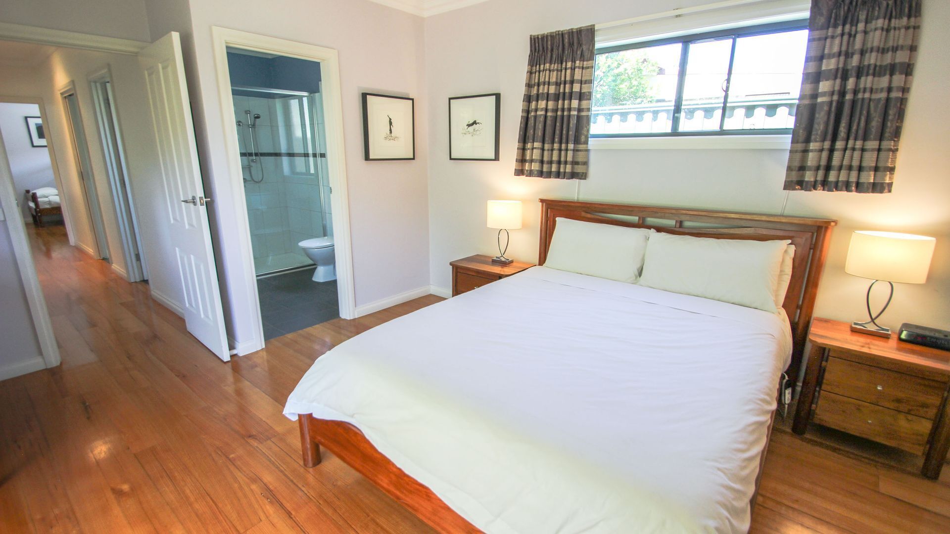 A bedroom with a large bed, two bedside tables with lamps, a window with curtains, a shower area, and framed pictures on the wall.