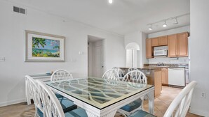 Un comedor con mesa de vidrio y sillas blancas, una cocina con muebles de madera y electrodomésticos blancos, y un cuadro enmarcado en la pared.