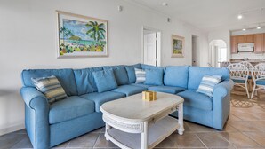 Una sala de estar con un sofá seccional azul, una mesa de centro blanca y un cuadro con una escena de playa en la pared.