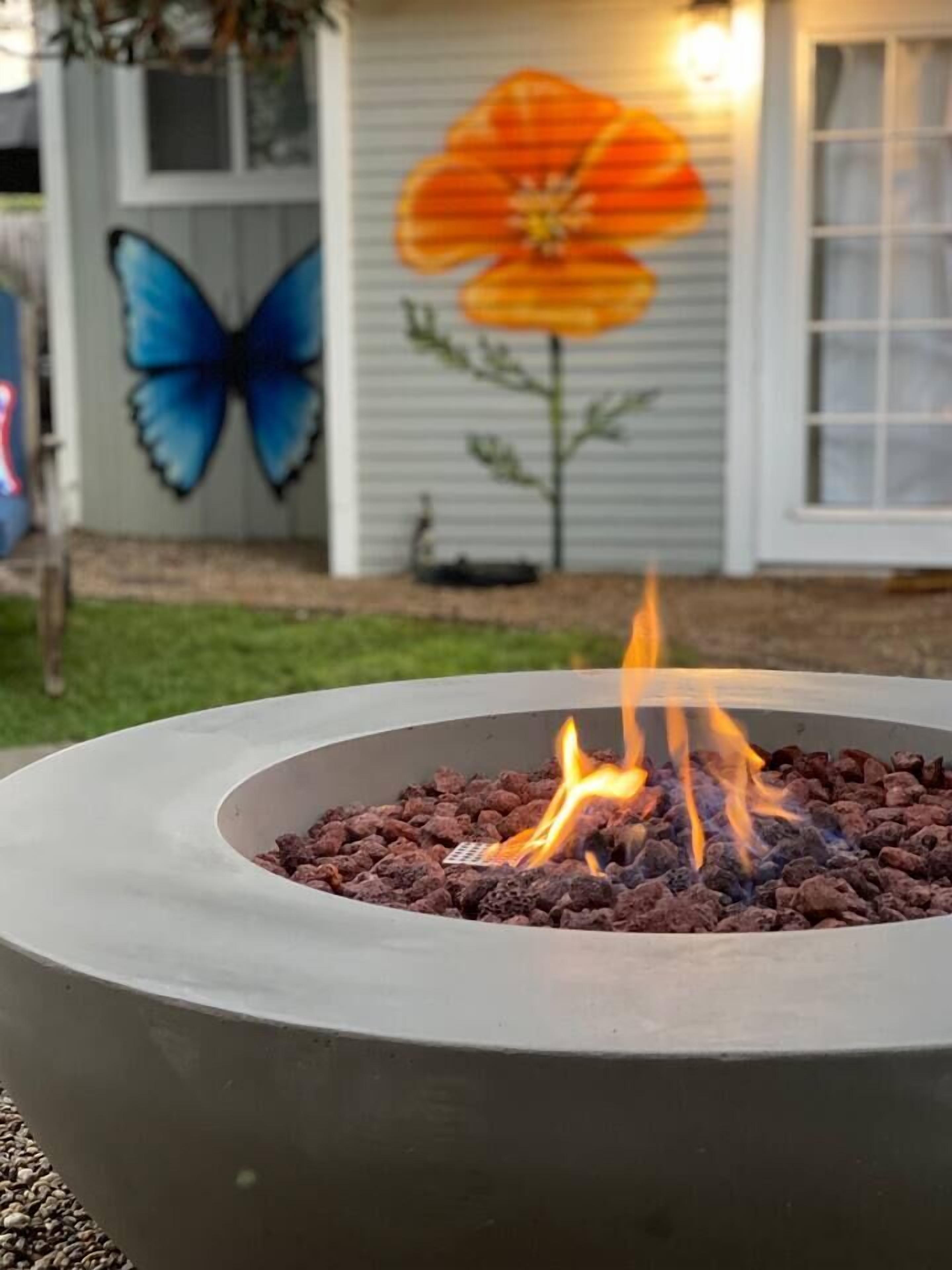 A fire pit with flames, surrounded by a concrete base, in front of a house with a painted flower and butterfly on the wall.